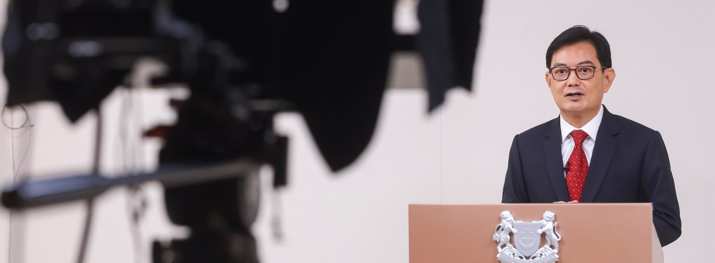 Lee Hsien Loong in suit behind a podium with the Singapore coat of arms, camera equipment visible.